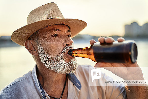 Älterer Mann trinkt bei Sonnenuntergang ein Bier am Flussufer