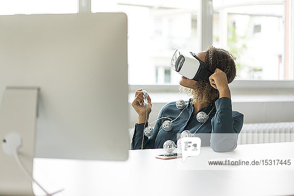 Young woman sitting at desk with chain of lights using Virtual Reality Glasses