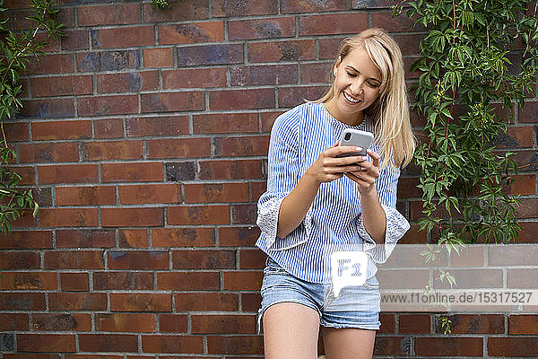 Lächelnde junge Frau steht mit einem Smartphone an einer Ziegelmauer