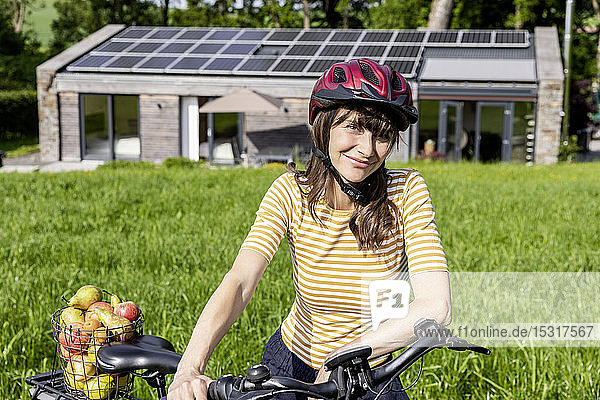 Porträt einer lächelnden Frau mit Fahrrad und Bio-Obst auf einer Wiese vor einem Haus