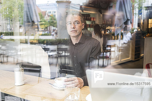 Man sitting in a cafe behind windowpane  drinking coffee with laptop