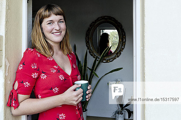 Porträt einer lächelnden jungen Frau mit Tasse Kaffee auf dem Balkon stehend
