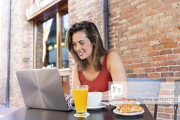 Lächelnder junger Blogger sitzt im Straßencafé und arbeitet am Laptop