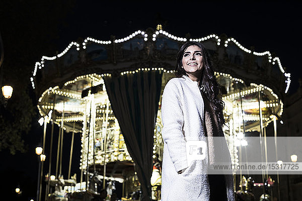 Porträt einer lächelnden jungen Frau  die nachts vor einem beleuchteten Karussell steht