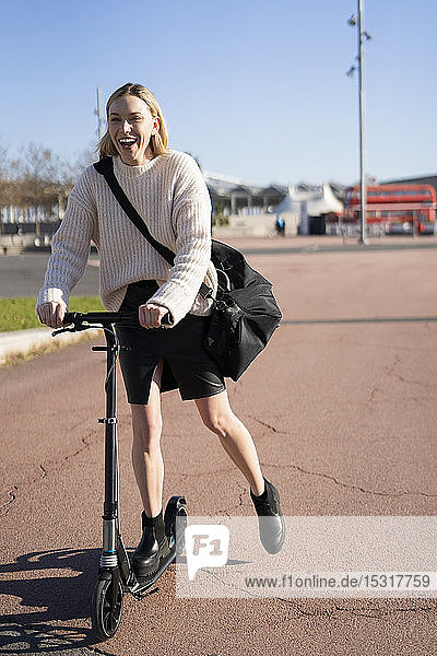 Lachende junge Frau mit Sporttasche auf Kickroller