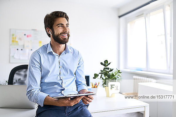 Lächelnder Geschäftsmann mit Kopfhörern  der ein digitales Tablet in seinem Büro benutzt
