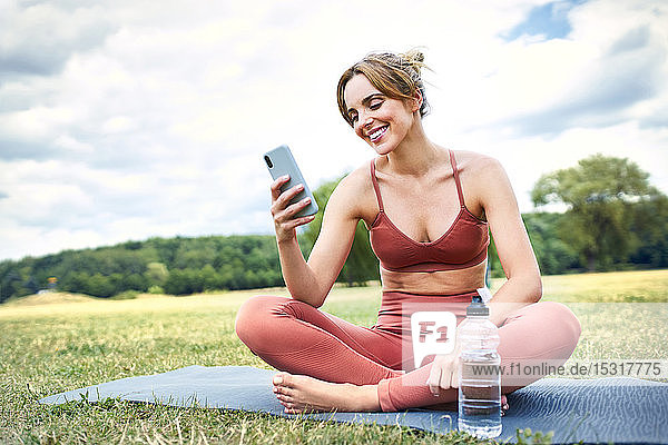Lächelnde Frau  die ein Smartphone benutzt  während sie eine Pause von der Yoga-Praxis im Freien einlegt