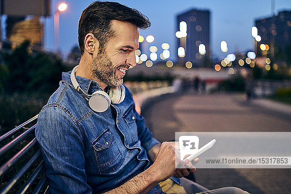 Lächelnder Mann benutzt sein Smartphone  während er abends auf einer Bank sitzt