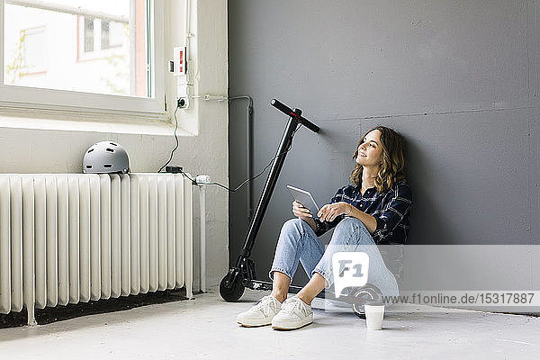 Young woman with e-scooter sitting on floor  using digital tablet