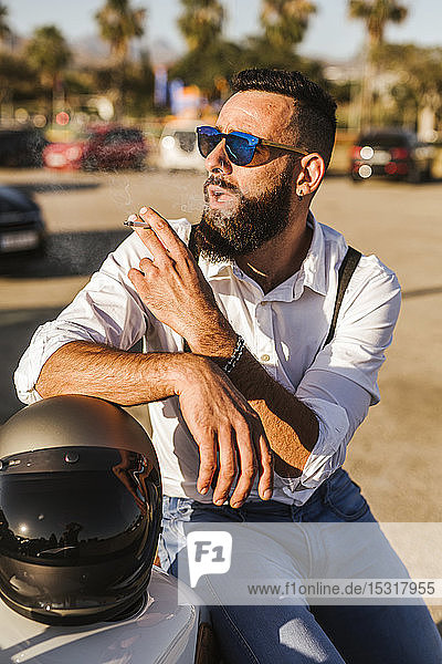 Portrait of bearded motorcyclist with mirrored sunglasses leaning on his helmet smoking