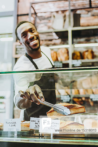 Lächelnder Mann arbeitet in einer Bäckerei und nimmt ein Stück Kuchen mit der Zange