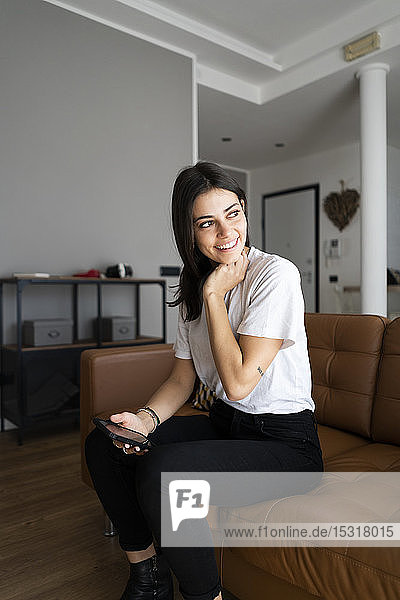 Lächelnde junge Frau auf der Couch zu Hause mit Handy