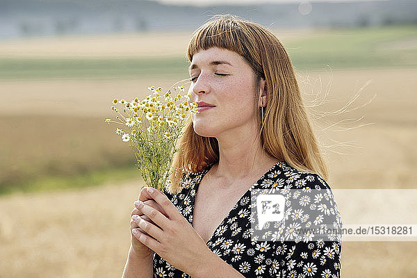 Lächelnde junge Frau mit geschlossenen Augen riecht in der Natur nach Kamillenblütensträußchen