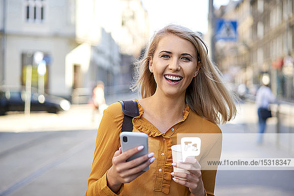 Porträt einer glücklichen jungen Frau mit Smartphone und Kaffee zum Mitnehmen in der Stadt