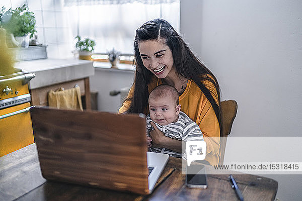 Lachende Mutter mit Baby mit Laptop auf dem Küchentisch