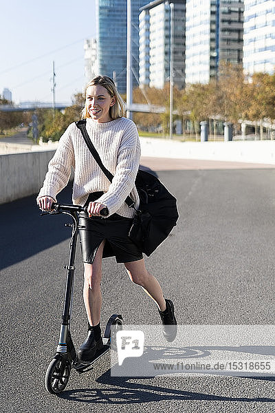 Smiling young woman with sports bag riding kick scooter
