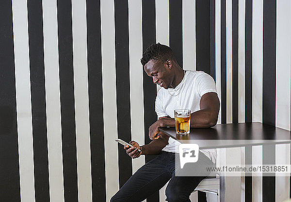 Junger Mann sitzt mit einem Getränk am Tisch und benutzt sein Handy