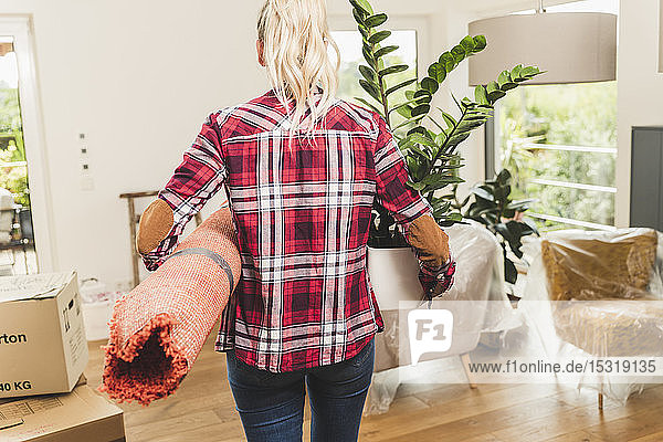 Rückansicht einer Frau  die mit Teppich und Topfpflanze in ihr neues Zuhause einzieht