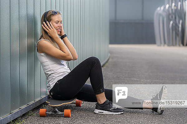 Smiling young woman sitting on longboard listening music with headphones