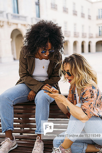 Multicultural happy women talking and using smartphone  sitting on bench