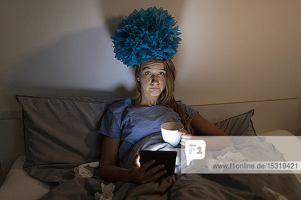 Ill young woman lying in bed at home with cup of tea at night using tablet
