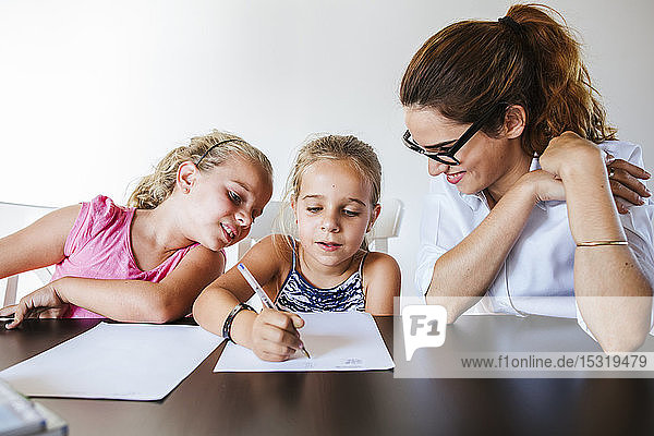 Lehrerin am Schreibtisch sitzend mit zwei Schülerinnen  die auf Papier schreiben