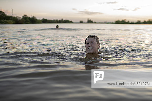 Porträt eines lächelnden Jungen  der bei Sonnenuntergang in einem See schwimmt