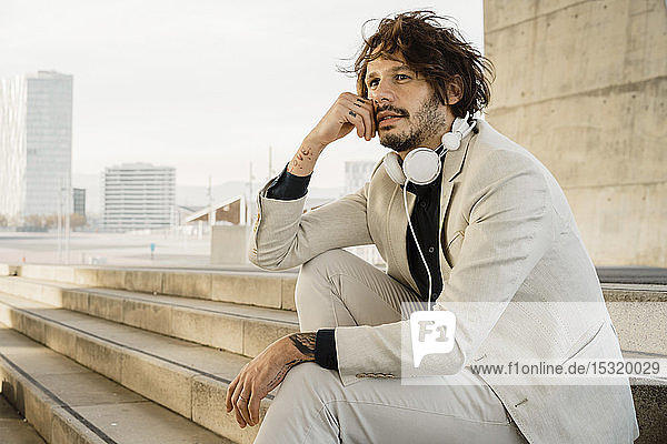 Porträt eines Geschäftsmannes mit Kopfhörern  der auf einer Treppe im Freien sitzt und in die Ferne schaut  Barcelona  Spanien
