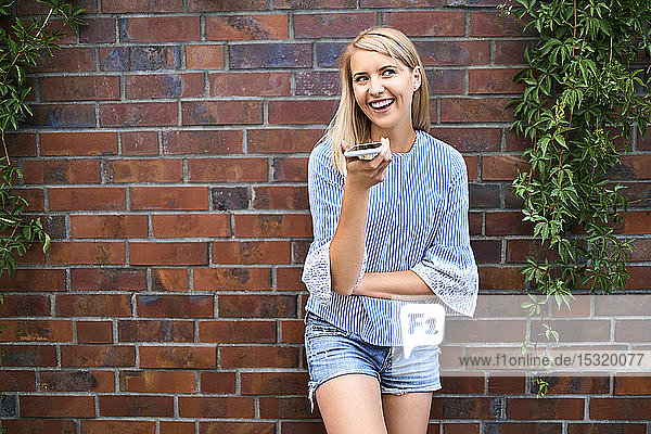 Happy young woman standing at a brick wall using smartphone