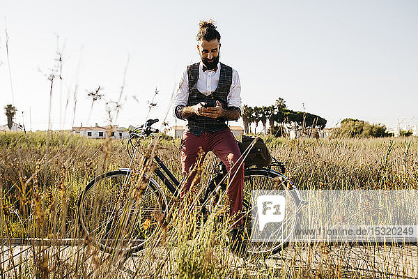 Gut gekleideter Mann mit seinem Fahrrad auf einem Holzsteg auf dem Land  der mit seinem Handy telefoniert