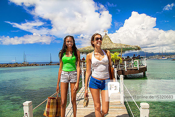 2 smiling girlfriends walking on a footbridge coming from a bar on stilts on a Caribbean beach in a paradisiacal landscape.