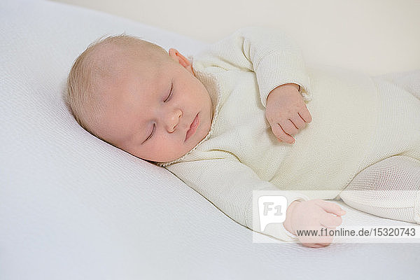 Junger Säugling in weißer Kleidung von 2 Monaten schläft auf einem weißen Bett.