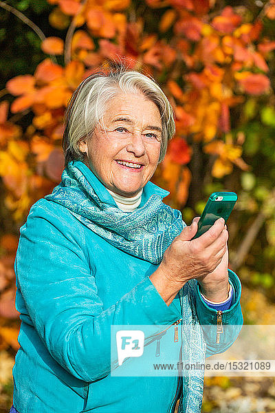 Sonniges Porträt einer lächelnden hübschen älteren Frau mit seinem Telefon vor einem Baum mit herbstlichen Farben.