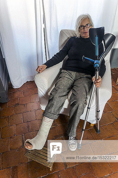 Frau auf einem Stuhl sitzend  mit einem eingegipsten Bein auf einer Fußstütze und mit Krücken in der Hand