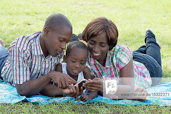 Diese Familie liegt auf einem Stück Stoff im Gras und konsultiert mit ihrem Kind ein Mobiltelefon.