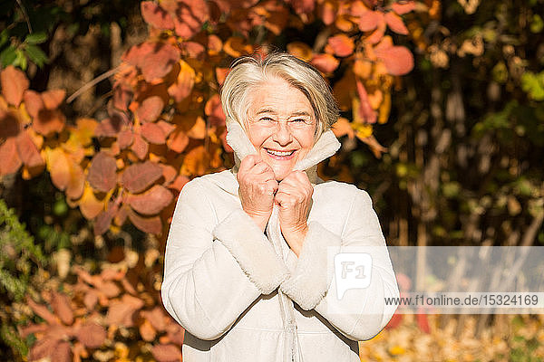 Sunny Porträt einer hübschen lächelnden Oma hält seinen Mantelkragen mit seinen Händen in der Natur in herbstlichen Farben.