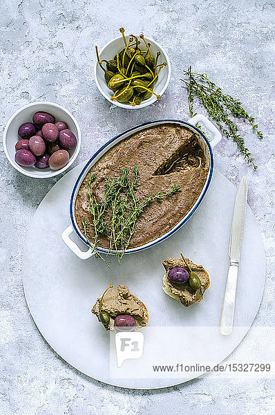 Leberpastete in Keramikform  dekoriert mit frischem Thymian und zwei Baguette-Sandwiches mit Pastete  Kapern  Oliven auf einem Marmorständer