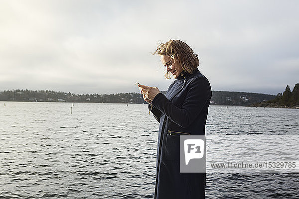 Young woman standing next to lake holding cell phone