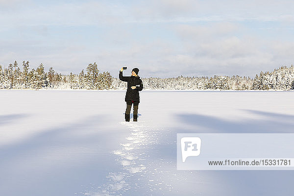 Frau fotografiert mit Smartphone am schneebedeckten Stora Skiren See in Lotorp, Schweden