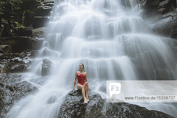 Woman wearing red swimsuit sitting by waterfall in Bali, Indonesia