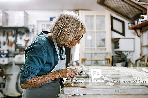 Seitenansicht eines älteren weiblichen Eigentümers  der Fensterrahmen auf der Werkbank in einer Werkstatt schabt