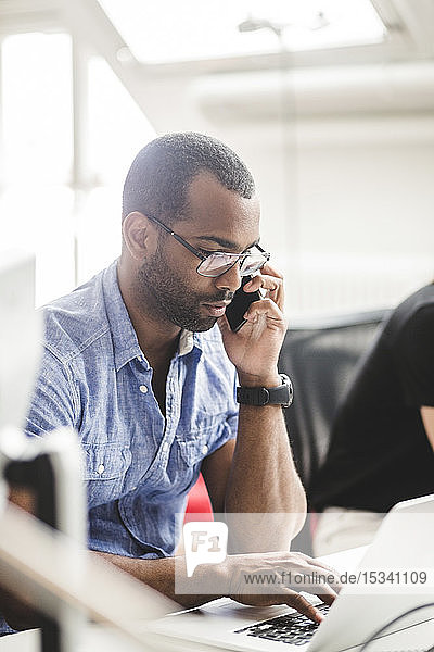 Businessman talking over smart phone while using laptop on desk in office