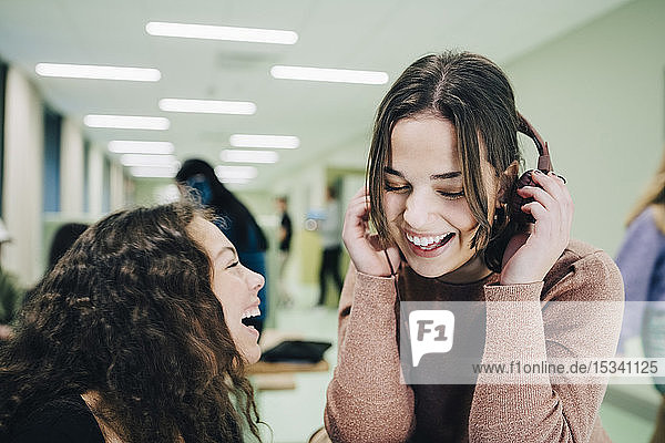 Fröhliches Teenager-Mädchen mit Kopfhörern sitzt mit einer Klassenkameradin im Flur der High School