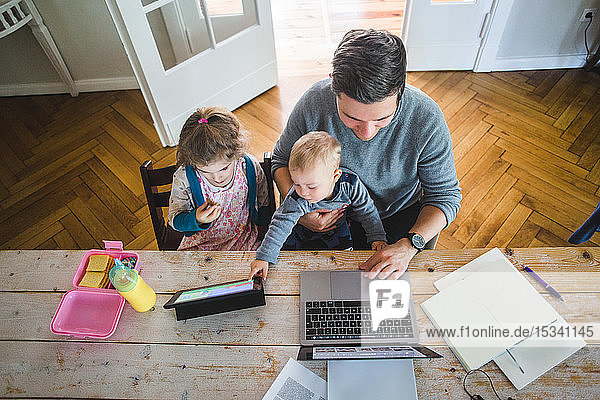 High angle view of man using laptop while sitting with cute son by daughter looking at digital tablet