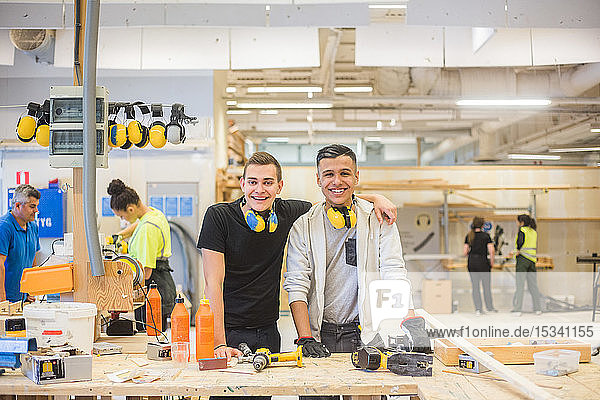 Portrait of confident young trainee standing with arm around teenage coworker at workbench at illuminated workplace