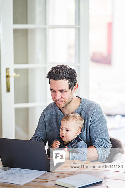 Mann arbeitet am Laptop  während er mit seinem Sohn am Tisch sitzt