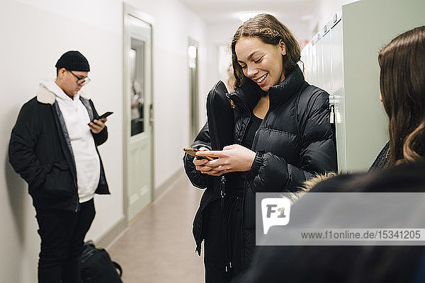 Smiling teenage student using smart phone in corridor