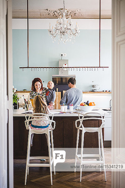 Lächelnde Frau  die einen Sohn trägt  während sie ein Mädchen ansieht  das zu Hause in der Küche lernt