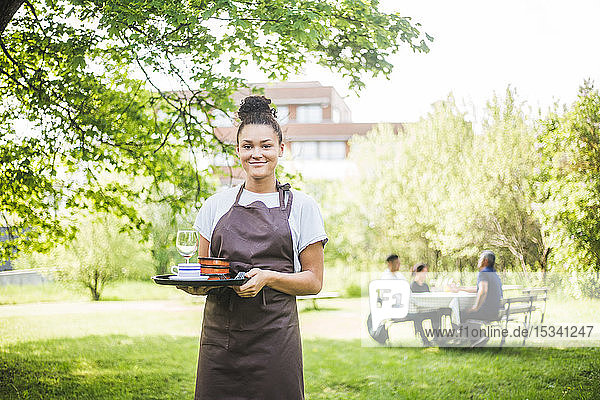 Porträt einer lächelnden jungen Kellnerin  die ein Serviertablett in einem Außencafé hält