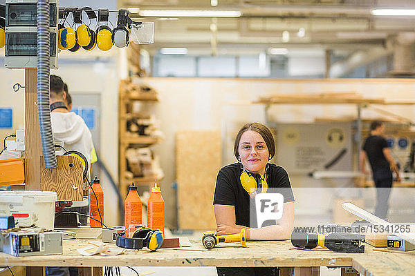 Portrait smiling young female carpentry trainee leaning on workbench with power tools at illuminated workshop